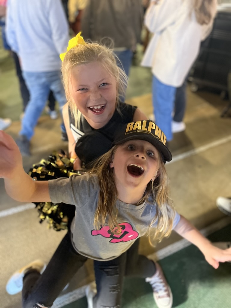 Two girls laughing at a Colorado Buffaloes game, one wearing a black Ralphie patch hat
