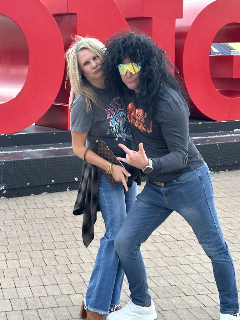 Couple in costume wigs and sunglasses posing at a Colorado concert venue sign